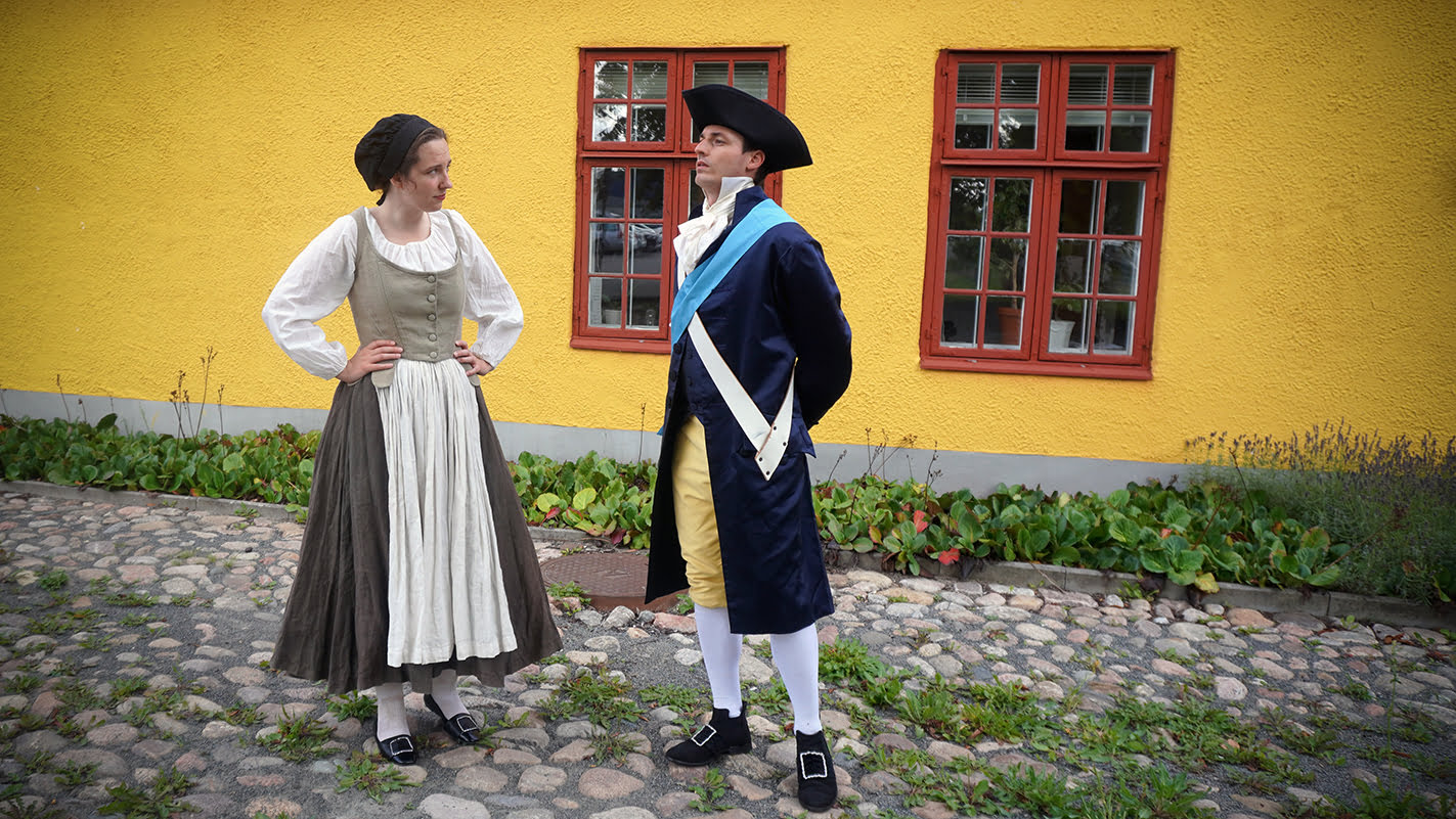 En kvinna i arbetarkläder och en man i militäruniform står mitt emot varandra framför ett hus.