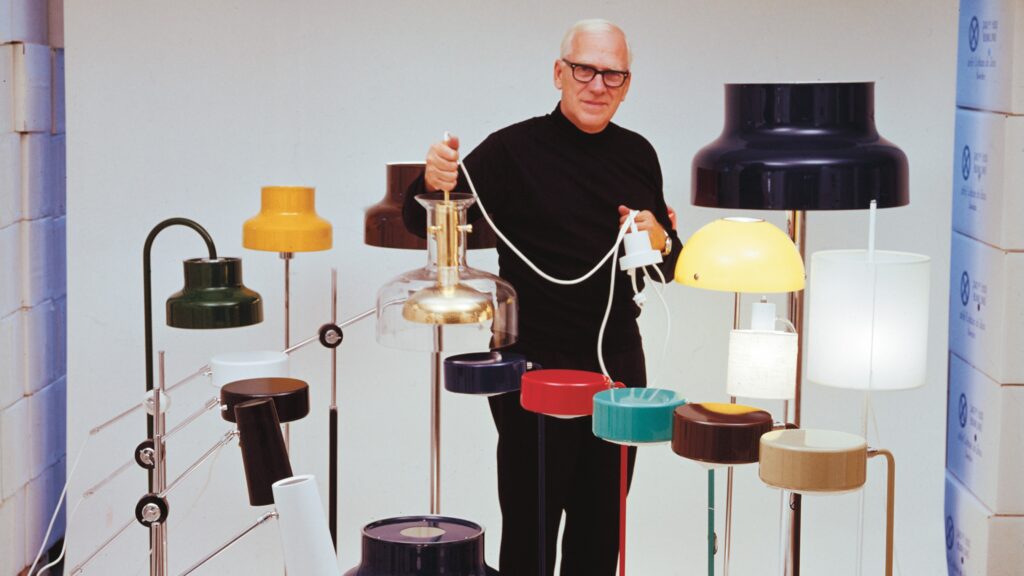En man står framför en grå bakgrund omringad av flera lampor och håller i en lampa.