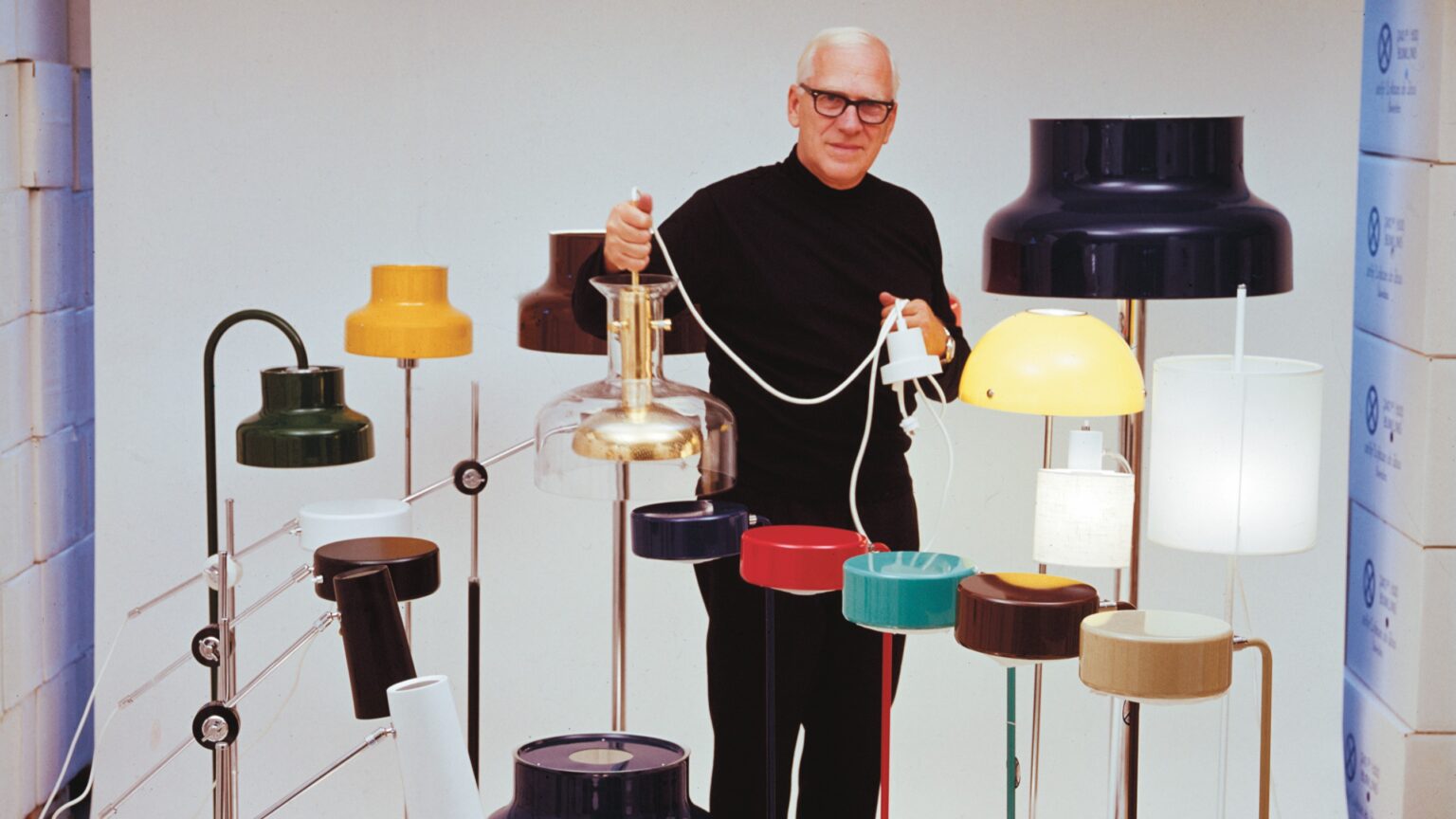 En man står framför en grå bakgrund omringad av flera lampor och håller i en lampa.