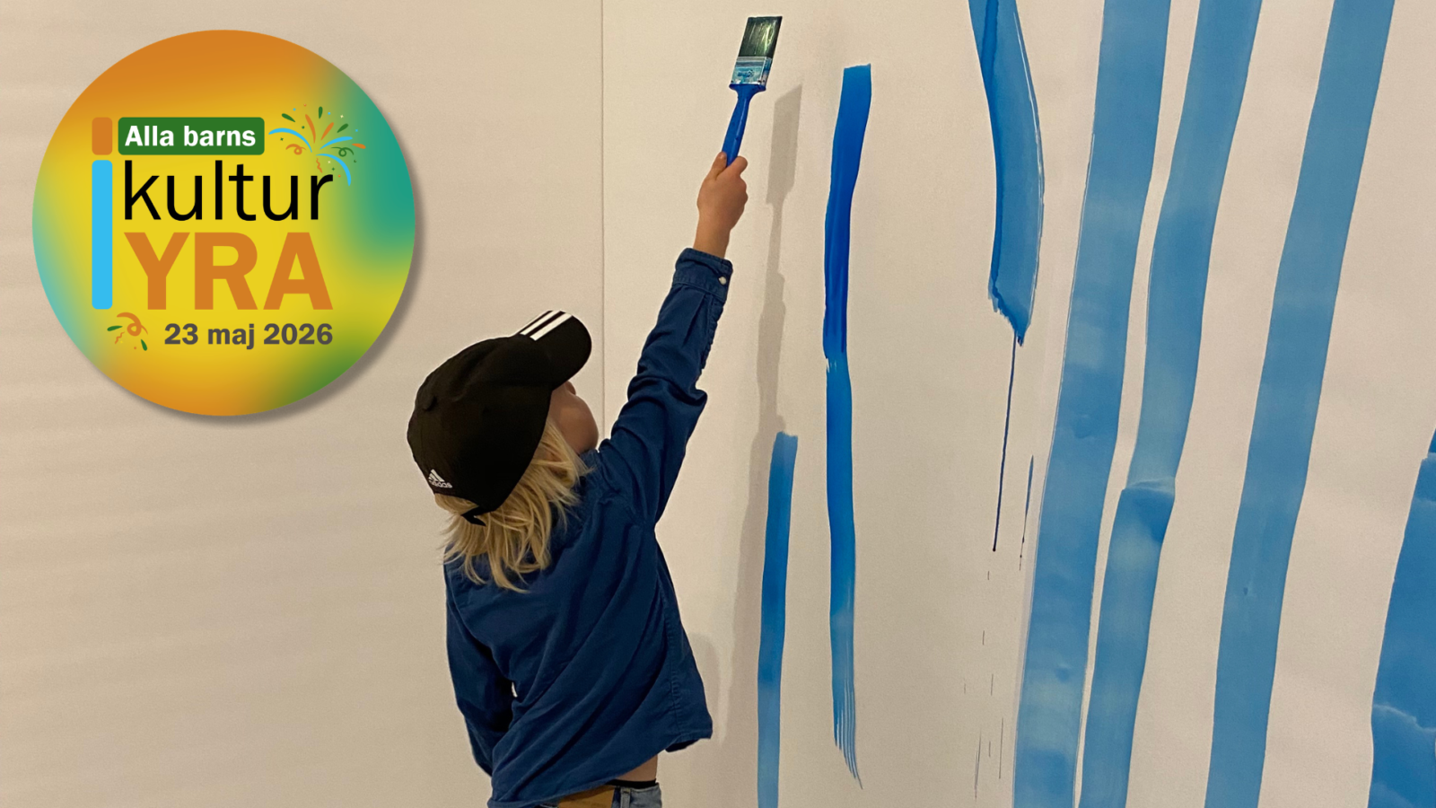 Ett barn i blå tröja och svart keps står med ryggen mot och målar med pensel blå färg på en vit vägg.