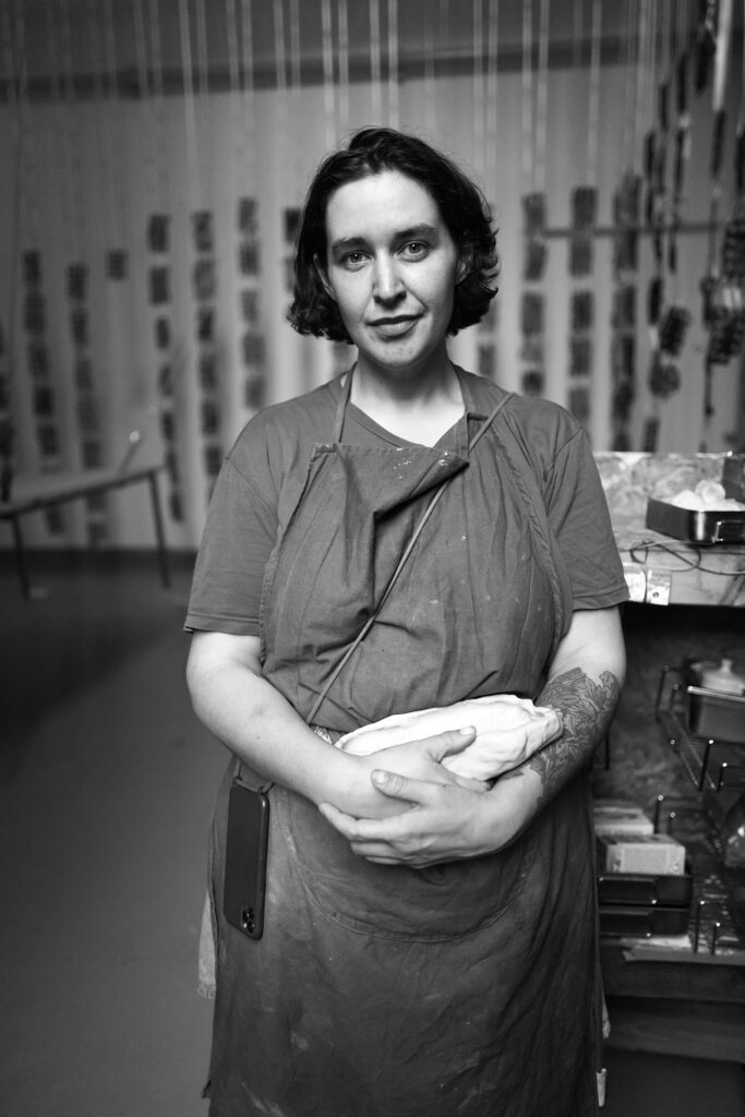 kvinna i ateljé med ett stycke lera i händerna som tittar in i kameran