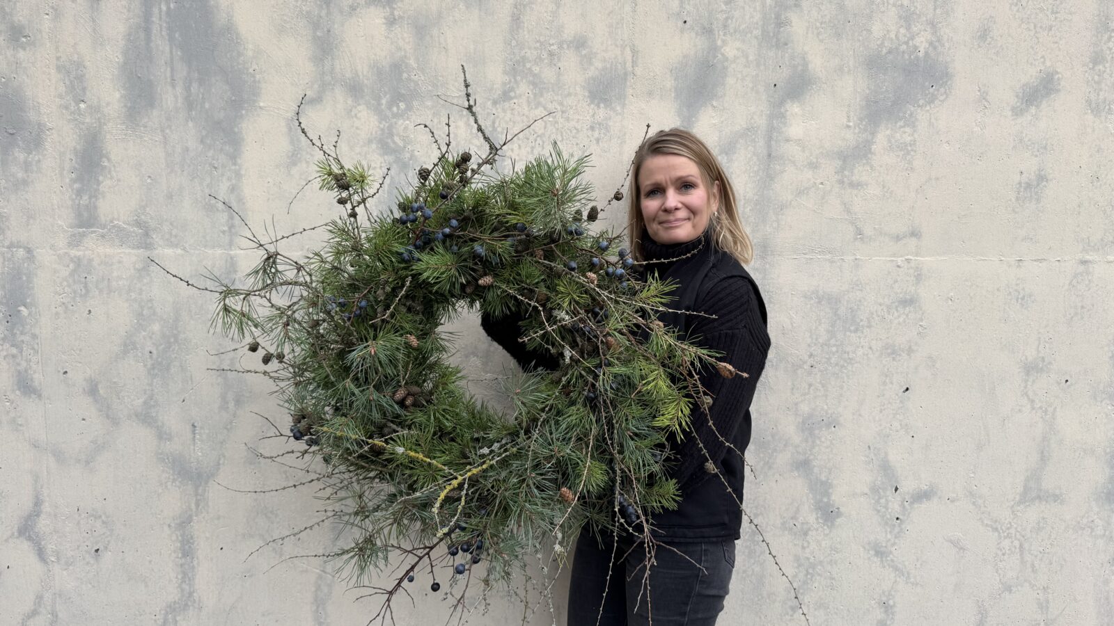 Kvinna står mot grå betongvägg och håller i en stor grön julkrans.